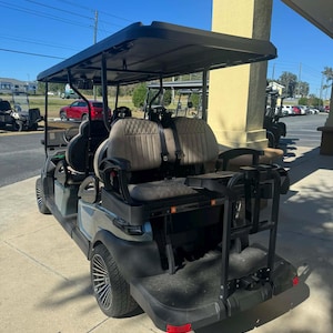 May include: A gray and black golf cart with a black roof and tan seats. The cart has a rear cargo area and is parked on a concrete surface. Other golf carts are visible in the background.