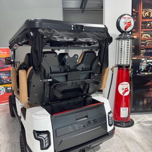 May include: A white and black Atlas golf cart featuring a rear storage compartment, tan seating, and a black roof. The vehicle has modern LED taillights and a hitch receiver. Beside it stands a vintage-style red Fire-Chief gasoline pump.