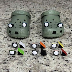 May include: Two olive green Crocs clogs with white and blue accents, and several colorful shoe charms. The charms include white circles, black, white, orange, green, and yellow accents. The clogs are on a speckled surface.