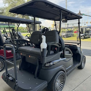 May include: A black golf cart with a black roof, seats, and wheels. The cart has a rear step and a white bottle holder. Other golf carts are visible in the background, including a red one.