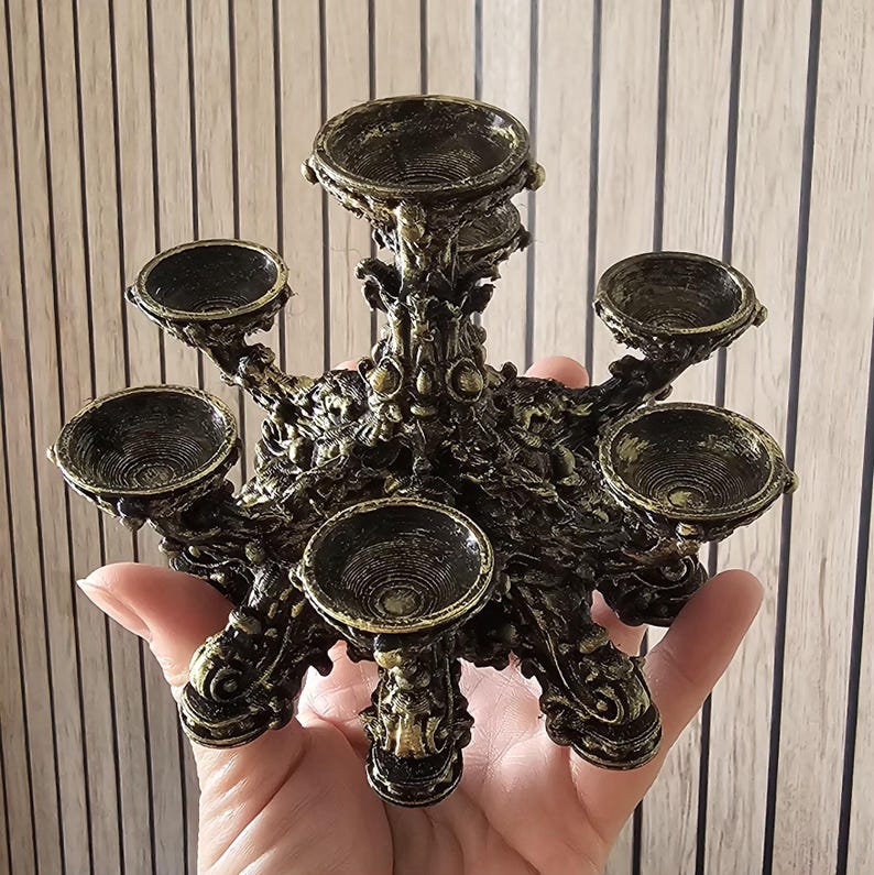 K&ouml;nnte beinhalten: Ein mehrstufiger Kerzenhalter mit einer zentralen, erh&ouml;hten Schale, die von f&uuml;nf tiefer gelegenen Schalen in kreisf&ouml;rmiger Anordnung umgeben ist. Das Objekt hat eine dunkel-schwarze Basis mit goldfarbenen Akzenten und aufwendigen, verzierten Details.