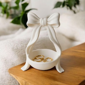 May include: A white ceramic ring dish in the shape of a bow. The dish features a small bowl for holding jewellery, with a bow at the top and ribbon-like sides. Two gold rings and a gold chain ring are inside the bowl. It rests on a wooden surface.