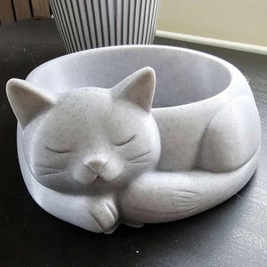 Könnte beinhalten: Ein grauer Katzen-Pflanztopf mit einem schlafenden Katzendesign. Der Pflanztopf ist rund mit einer strukturierten Oberfläche und einer kleinen Öffnung für Pflanzen. Ein schwarz-weiß gestreifter Topf befindet sich im Hintergrund.