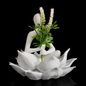 May include: A white speckled figurine of a person sitting in a lotus position, holding a bouquet of green leaves and white flowers. The figure is nestled within a large, open white speckled lotus flower base against a solid black background.