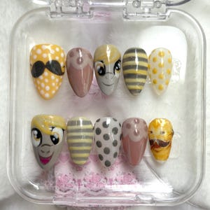 May include: A set of ten artificial nails with various designs. The nails feature cartoon character faces, polka dots, stripes, and a mustache. The color palette includes yellow, gray, brown, and pink, all encased in a clear plastic container.