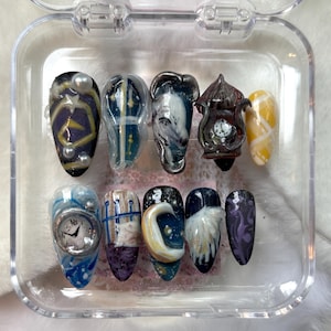 Peut inclure: Un ensemble de dix faux ongles avec divers motifs. Les ongles présentent des œuvres d'art détaillées, notamment des thèmes célestes, des horloges et des motifs abstraits. Les couleurs incluent le bleu, le jaune, le violet et le blanc, certains ongles ayant une finition brillante.