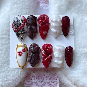 May include: A set of ten artificial nails in various designs. The nails feature a mix of colors, including white, red, and gold, with decorative elements such as floral patterns, sculpted figures, and lip art. Some nails have a glossy finish.