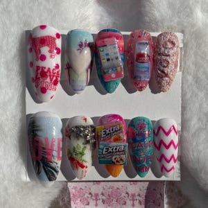 Könnte beinhalten: Ein Set aus zehn künstlichen Nägeln mit verschiedenen Designs. Die Nägel weisen eine Vielzahl von Farben auf, darunter Rosa, Blau und Weiß, und enthalten Bilder von einem Telefon, Kaugummi und Text. Einige Nägel haben Glitzerakzente und Text, der "PEACE LOVE PINK" und "LOVE" lautet.