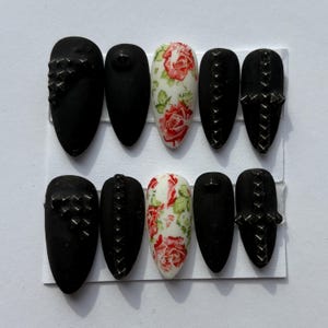 Könnte beinhalten: Ein Set aus zehn künstlichen Nägeln in Schwarz und floralen Designs. Die schwarzen Nägel haben matte Oberflächen, Nieten und Kreuzmuster. Die floralen Nägel haben rote Rosen und grüne Blätter auf weißem Grund.