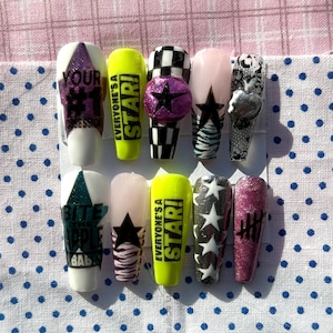 Puede incluir: Un conjunto de diez uñas postizas con diversos diseños. Las uñas presentan texto, estrellas, patrones de cuadros y purpurina. Los colores incluyen blanco, negro, rosa, verde y amarillo. Algunas uñas tienen el texto "EVERYONE'S A STAR!"
