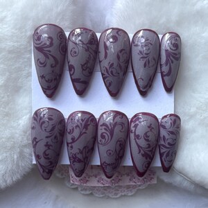 May include: Set of ten almond-shaped false nails. Each nail is painted with a light purple base and a dark purple floral design. The nails are arranged on a white card, with a soft, white, textured background.