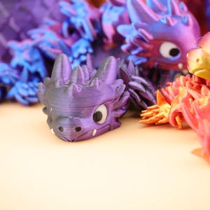 以下が含まれることがあります： カラフルな3Dプリントのドラゴンフィギュアのコレクション。ドラゴンは紫、青、赤、オレンジなど、さまざまな色を特徴としています。各ドラゴンは、スパイクと大きな目を持つ詳細なデザインで、ファンタジーテーマを作成しています。