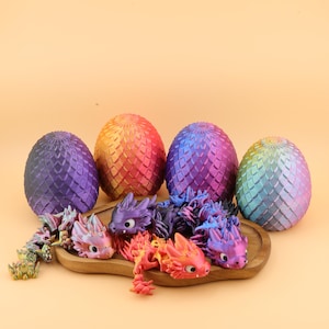 Könnte beinhalten: Eine Sammlung farbenfroher Dracheneier und beweglicher Drachenfiguren. Die Eier sind in den Farbtönen Lila, Orange, Rot, Blau und Grün gehalten. Die Drachen sind in verschiedenen Farben gehalten und werden auf einem Holztablett präsentiert.