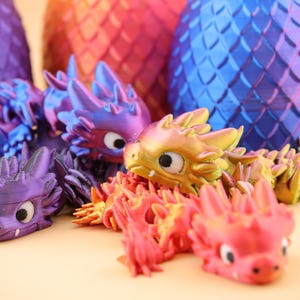 以下が含まれることがあります： カラフルな3Dプリントのドラゴンフィギュアとドラゴンの卵の彫刻のコレクション。ドラゴンは紫、金、赤の色合いです。卵はオレンジ、赤、青の色合いです。