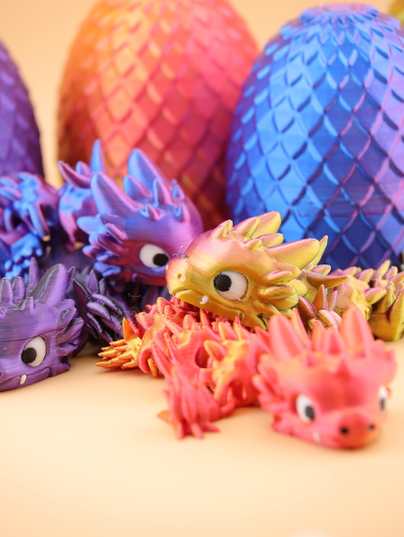 以下が含まれることがあります： カラフルな3Dプリントのドラゴンフィギュアとドラゴンの卵の彫刻のコレクション。ドラゴンは紫、金、赤の色合いです。卵はオレンジ、赤、青の色合いです。