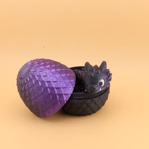 以下が含まれることがあります： 紫と黒のドラゴンの卵と、中にドラゴンの置物。 卵は開いており、ドラゴンの頭と体の一部が見えています。 卵はダイヤモンドのような模様のテクスチャです。