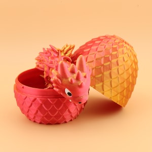 以下が含まれることがあります： お揃いの卵から出てくる3Dプリントのドラゴン。ドラゴンと卵は主に赤色で、金色のアクセントが付いています。卵は開いており、ドラゴンの頭と体の一部が見えます。卵の殻は、鱗のようなテクスチャパターンになっています。