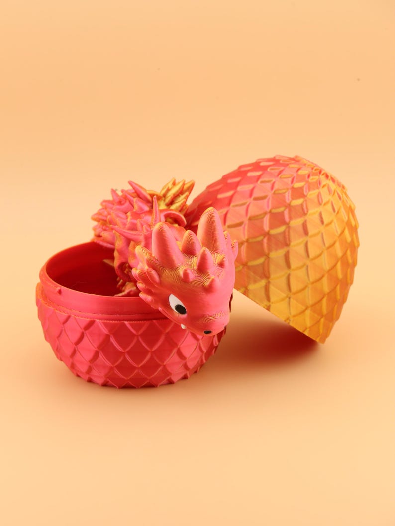 以下が含まれることがあります： お揃いの卵から出てくる3Dプリントのドラゴン。ドラゴンと卵は主に赤色で、金色のアクセントが付いています。卵は開いており、ドラゴンの頭と体の一部が見えます。卵の殻は、鱗のようなテクスチャパターンになっています。