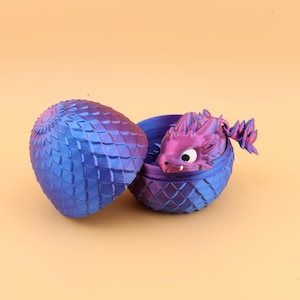 以下が含まれることがあります： 青と紫のドラゴンの卵。3Dプリントされており、鱗のようなテクスチャパターンがあります。卵は開いており、白い目をした小さな紫色のドラゴンのフィギュアが現れています。ドラゴンは卵から出てきており、ファンタジーをテーマにしたアイテムです。