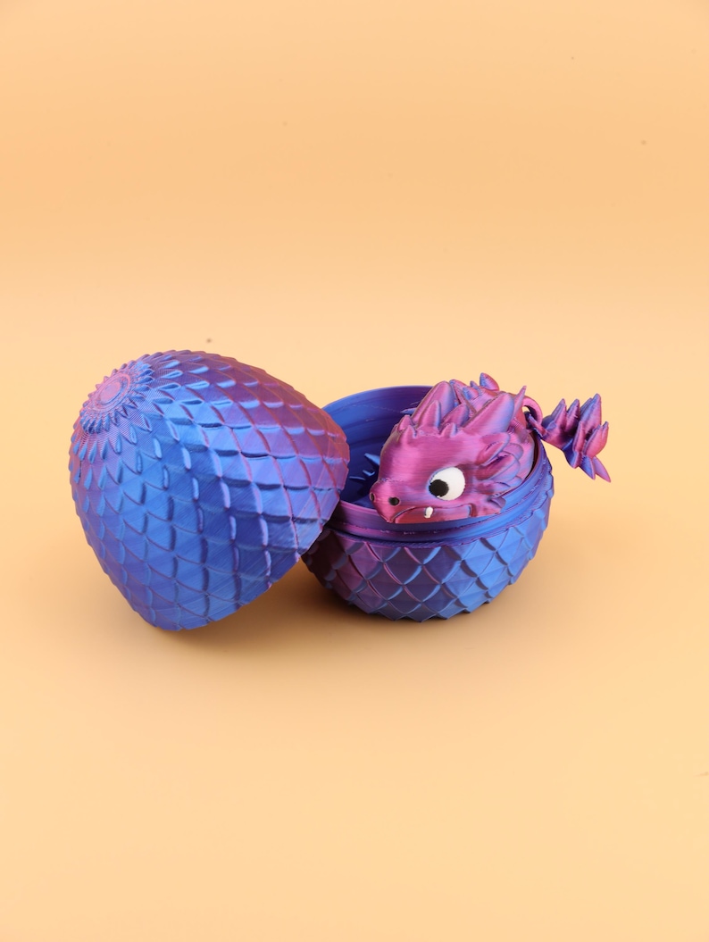 以下が含まれることがあります： 青と紫のドラゴンの卵。3Dプリントされており、鱗のようなテクスチャパターンがあります。卵は開いており、白い目をした小さな紫色のドラゴンのフィギュアが現れています。ドラゴンは卵から出てきており、ファンタジーをテーマにしたアイテムです。