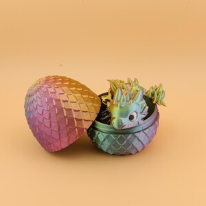 以下が含まれることがあります： 小さなドラゴンフィギュアが現れる、開いた装飾的な卵。卵とドラゴンは虹色で、金色から紫、緑へと色が変化する。卵は鱗のような模様のテクスチャを持っています。