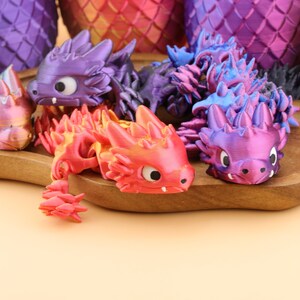 以下が含まれることがあります： 紫、赤、オレンジ、青の色合いの、カラフルな3Dプリントのドラゴンフィギュアのコレクション。ドラゴンは詳細な鱗と大きな目を持っています。背景には、さらにドラゴンスケール模様のオブジェクトがあります。