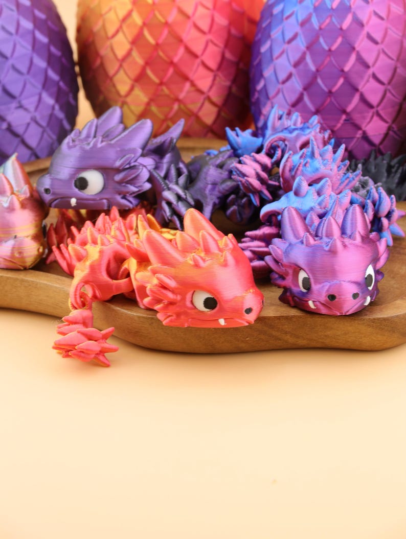 以下が含まれることがあります： 紫、赤、オレンジ、青の色合いの、カラフルな3Dプリントのドラゴンフィギュアのコレクション。ドラゴンは詳細な鱗と大きな目を持っています。背景には、さらにドラゴンスケール模様のオブジェクトがあります。
