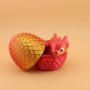 以下が含まれることがあります： 装飾的な卵から孵化する3Dプリントのドラゴン。卵は2色で、金色の黄色い上部と赤いベースがあります。ドラゴンは赤色で黄色のアクセントがあり、卵から出ています。