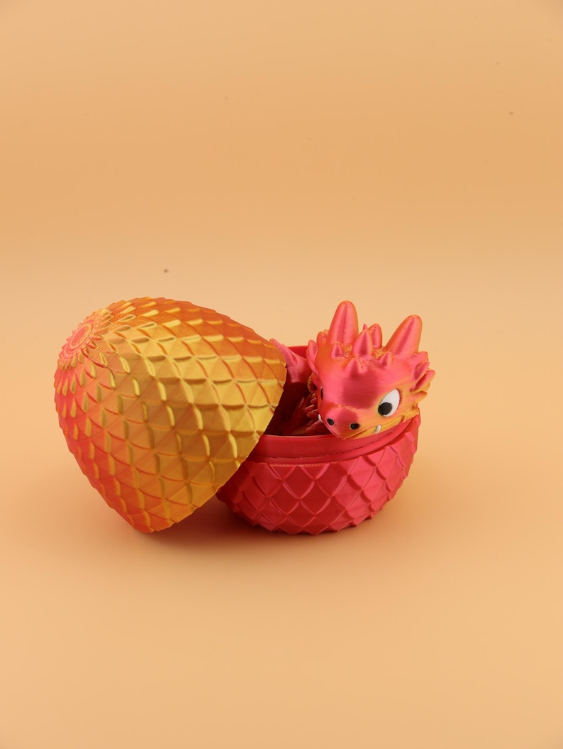 以下が含まれることがあります： 装飾的な卵から孵化する3Dプリントのドラゴン。卵は2色で、金色の黄色い上部と赤いベースがあります。ドラゴンは赤色で黄色のアクセントがあり、卵から出ています。
