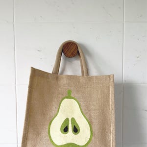 May include: A brown jute tote bag with a green pear design. The pear is cut in half and has a white interior with black seeds.