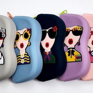 May include: A collection of five colorful eyeglass cases, each featuring a stylized woman's face with oversized sunglasses. The cases are in shades of gray, blue, black, purple, and pink, with red zipper pulls.