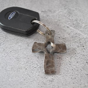 May include: A black Ford car key attached to a silver-toned metal cross keychain. The cross features a hammered, textured surface with a small diamond-shaped cutout in the center, connected to the key fob by a circular metal split ring.