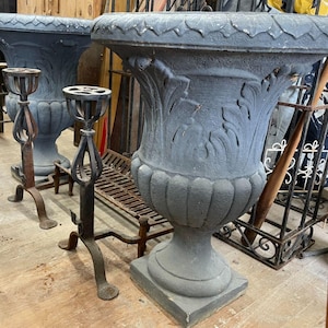 May include: Two large, ornate, gray urn planters with detailed floral relief patterns. The planters are on square bases. Two wrought iron stands are in front of the planters. The stands have a decorative design and are made of dark metal.