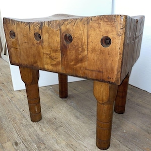 May include: A large, antique butcher block table made of wood. The table has a rectangular top with a curved indentation and four sturdy, cylindrical legs. The wood has a warm, brown tone with visible grain and wear.