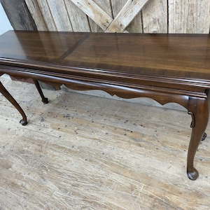 May include: A dark brown wooden console table with a rectangular top and curved legs. The table has a scalloped apron and a glossy finish. The table is approximately 60 inches long and 18 inches wide.