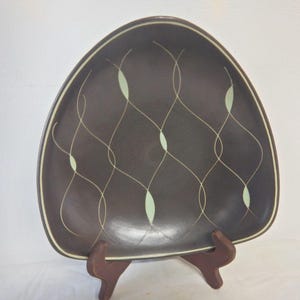 May include: A brown, triangular-shaped plate with a cream-colored border and a decorative pattern of intersecting lines and green ovals. The plate is displayed on a wooden stand, showcasing its mid-century modern design.