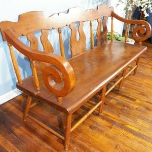 May include: A wooden bench with a curved back and scrolled armrests. The bench is made of light brown wood and has a long seat. The back features decorative cutouts and vertical spindles. The bench is supported by four legs and horizontal supports.