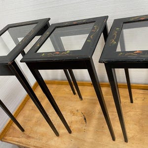 May include: Three black display tables with glass tops and slender legs. The edges of the tables are decorated with colorful floral and leaf designs. The tables are arranged on a light-colored wooden surface.