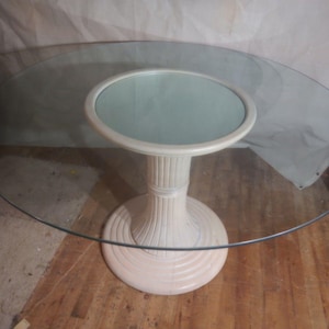 May include: A round glass-top table with a cream-colored base. The base has a pedestal design with a circular top and bottom. The glass top is oval-shaped. The table is set on a wooden floor.