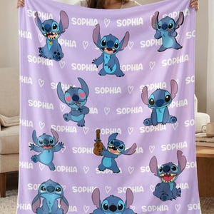 May include: A lavender blanket featuring multiple illustrations of a blue cartoon character. The word "SOPHIA" and heart symbols are printed across the blanket. The blanket is held up by a person.