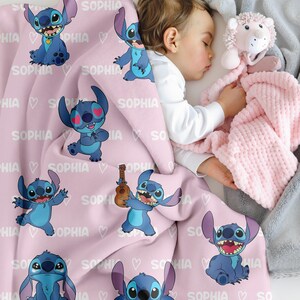 May include: A pink baby blanket featuring multiple cartoon images of a blue alien character. The blanket has the name "SOPHIA" printed on it, along with white hearts. A sleeping baby is partially visible.
