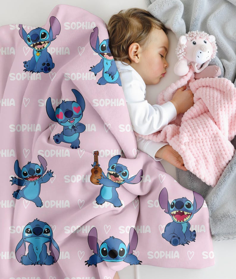May include: A pink baby blanket featuring multiple cartoon images of a blue alien character. The blanket has the name "SOPHIA" printed on it, along with white hearts. A sleeping baby is partially visible.