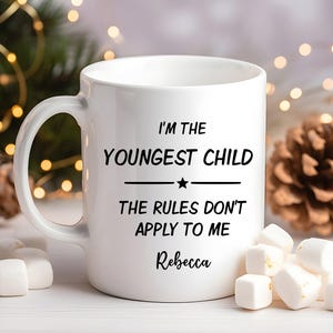 May include: White ceramic mug with the text "I'M THE YOUNGEST CHILD THE RULES DON'T APPLY TO ME" and the name "Rebecca". The mug is surrounded by marshmallows and festive decorations.