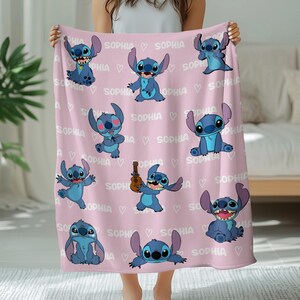 May include: A pink blanket featuring multiple blue cartoon character illustrations. The word "SOPHIA" is repeated throughout the design, along with small white hearts. The blanket is held up by a person.