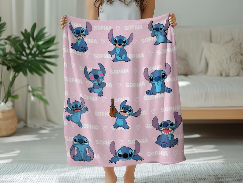 May include: A pink blanket featuring multiple blue cartoon character illustrations. The word "SOPHIA" is repeated throughout the design, along with small white hearts. The blanket is held up by a person.
