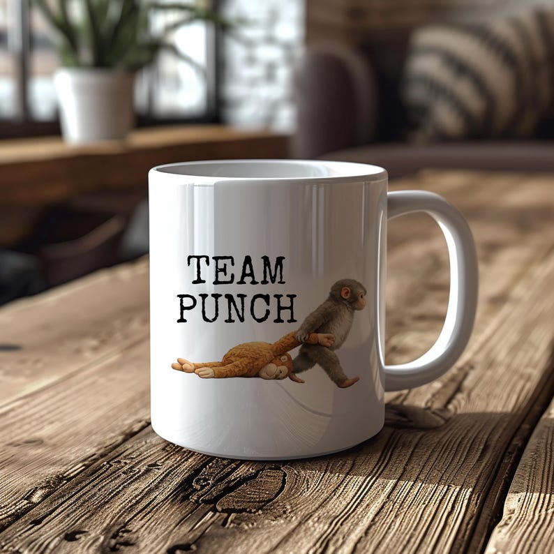 May include: White ceramic mug with the text "TEAM PUNCH" and a cartoon illustration of two monkeys. One monkey is pulling the other by the arm. The mug is sitting on a wooden surface.