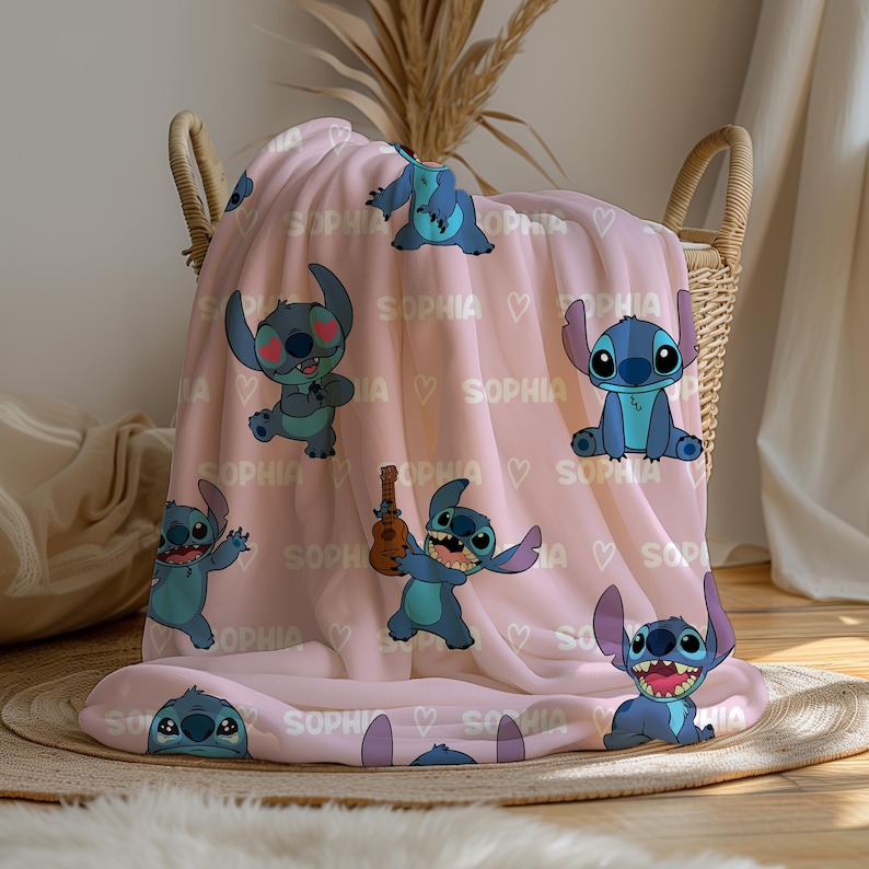 May include: A light pink fleece blanket featuring multiple cartoon images of Stitch, with the name "SOPHIA" and hearts printed across the blanket. The blanket is folded in a wicker basket.