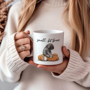 May include: White ceramic mug with the text "small, but brave" in black script. The mug features a black and gray illustration of a small ape sitting on a brown stuffed animal. The mug is held by a person wearing a white sweater.