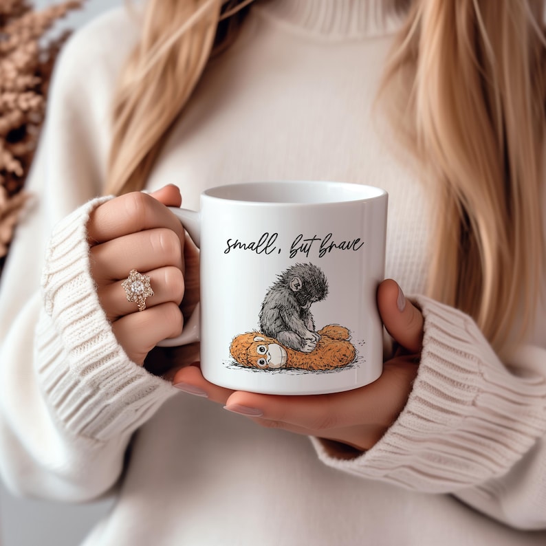 May include: White ceramic mug with the text "small, but brave" in black script. The mug features a black and gray illustration of a small ape sitting on a brown stuffed animal. The mug is held by a person wearing a white sweater.