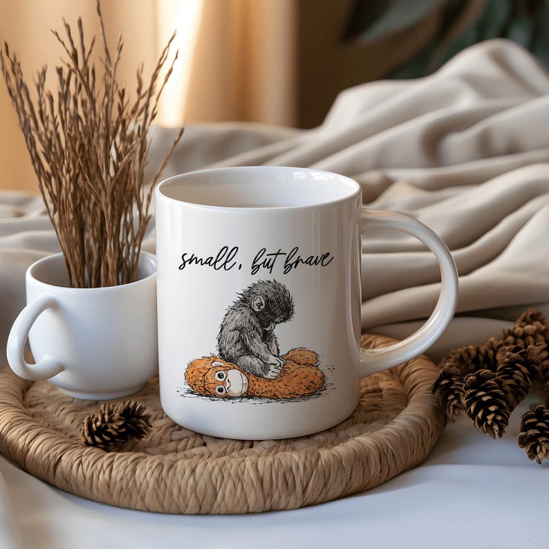 May include: White ceramic mug with a whimsical illustration of a small, gray creature atop a brown one, with the words "small, but brave" in black script. The mug is on a woven tray with a small white cup and dried plants.